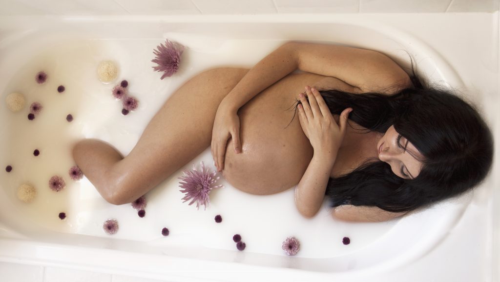 Pregnant woman holding tummy lying in a milk bath with flowers