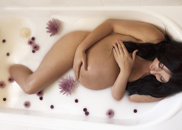 Pregnant woman holding tummy lying in a milk bath with flowers