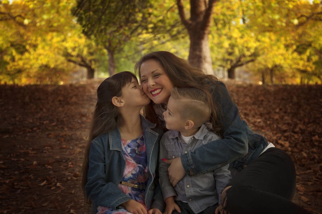 kids kissing mother sitting in park with autumn leaves